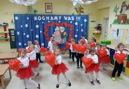 Dzieci z grupy Kubusie Puchatki (Gołębin Stary) w odświętnych strojach podczas tańca z czerwonymi balonami w kształcie serca dla babci i dziadka na tle gazetki z napisem  (photo)